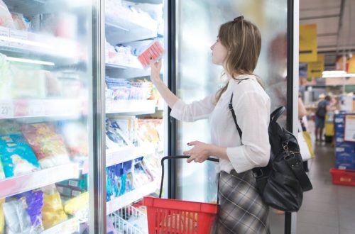 A woman opening a freezer door at the grocery store and selecting a frozen food item. She's holding a basket.