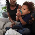 A little black toddler boy in a blue polo shirt sitting with a black woman with braids on a couch, breathing into an inhaler.