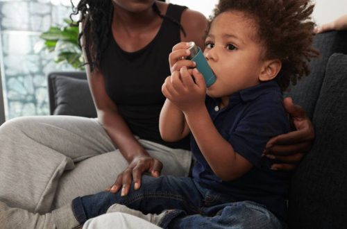 A little black toddler boy in a blue polo shirt sitting with a black woman with braids on a couch, breathing into an inhaler.