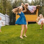 A woman in a denim dress swinging a small toddler around with other children running around them in the backyard.