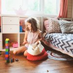 A young girl puts her toys away in a red and white knitted bag on her bedroom floor. There's a set of shelves behind her.