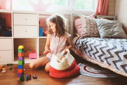 A young girl puts her toys away in a red and white knitted bag on her bedroom floor. There's a set of shelves behind her.