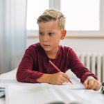 A young blond-haired blue-eyed boy in a red shirt sitting at a desk with notebooks and a colored pencil with an open laptop.