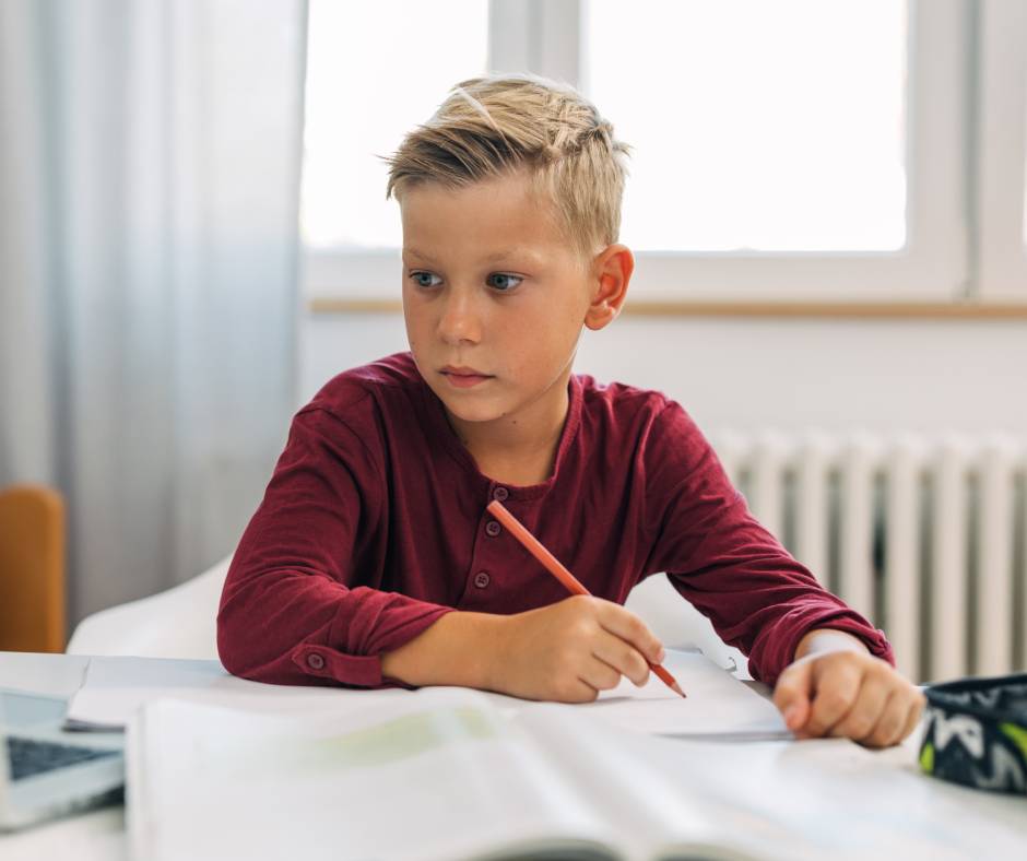 A young blond-haired blue-eyed boy in a red shirt sitting at a desk with notebooks and a colored pencil with an open laptop.