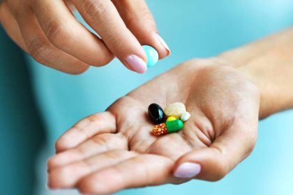 A pair of hands with pink nails and a blue background. One lifts a pill out of a pile in the other’s palm.