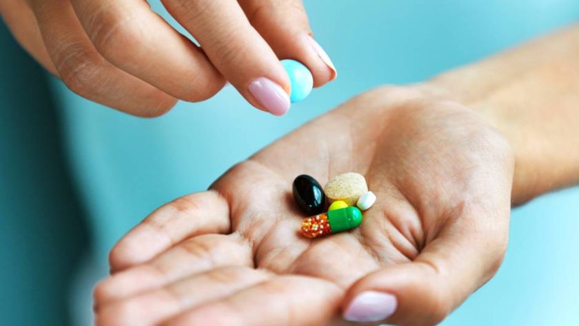 A pair of hands with pink nails and a blue background. One lifts a pill out of a pile in the other’s palm.