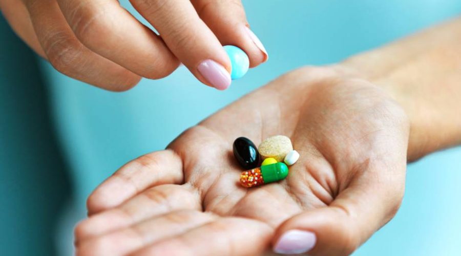 A pair of hands with pink nails and a blue background. One lifts a pill out of a pile in the other’s palm.