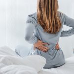 A woman sits on the edge of her bed and holds her lower back in pain. She is wearing a grey pajama set.