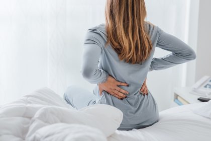 A woman sits on the edge of her bed and holds her lower back in pain. She is wearing a grey pajama set.