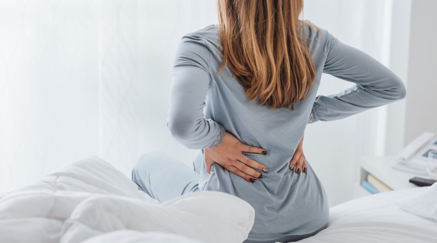 A woman sits on the edge of her bed and holds her lower back in pain. She is wearing a grey pajama set.