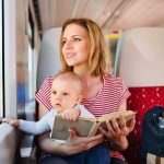 A woman in a striped shirt sits on a train seat holding a baby and a book, with a blue bag and a teddy bear beside her.