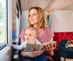 A woman in a striped shirt sits on a train seat holding a baby and a book, with a blue bag and a teddy bear beside her.