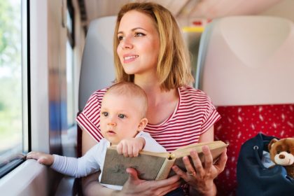 A woman in a striped shirt sits on a train seat holding a baby and a book, with a blue bag and a teddy bear beside her.