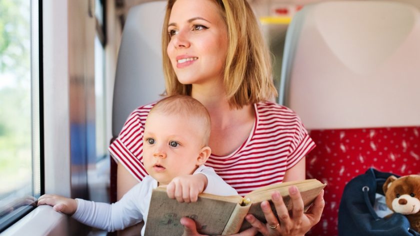 A woman in a striped shirt sits on a train seat holding a baby and a book, with a blue bag and a teddy bear beside her.