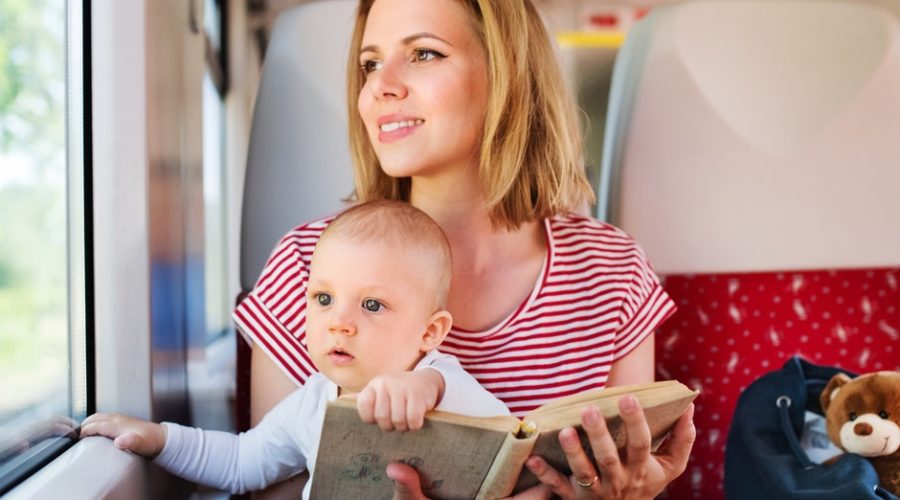 A woman in a striped shirt sits on a train seat holding a baby and a book, with a blue bag and a teddy bear beside her.