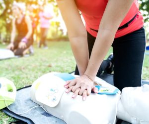 A close-up showing someone wearing an orange shirt practicing chest compressions on a dummy while outside on the grass.