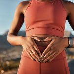 A woman in fitness apparel holding her hands over her stomach while wearing a watch and mountain ridges behind her.