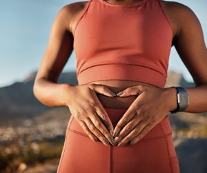 A woman in fitness apparel holding her hands over her stomach while wearing a watch and mountain ridges behind her.