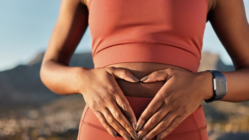 A woman in fitness apparel holding her hands over her stomach while wearing a watch and mountain ridges behind her.