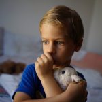 A young boy wearing a blue t-shirt holds a plush, white bunny against his chest while he sucks his thumb.