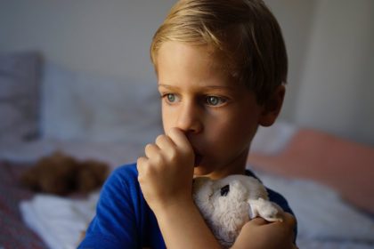 A young boy wearing a blue t-shirt holds a plush, white bunny against his chest while he sucks his thumb.