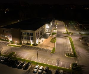An office building parking lot set at night. While the entrance is empty, there are some cars parked in a nearby lot.