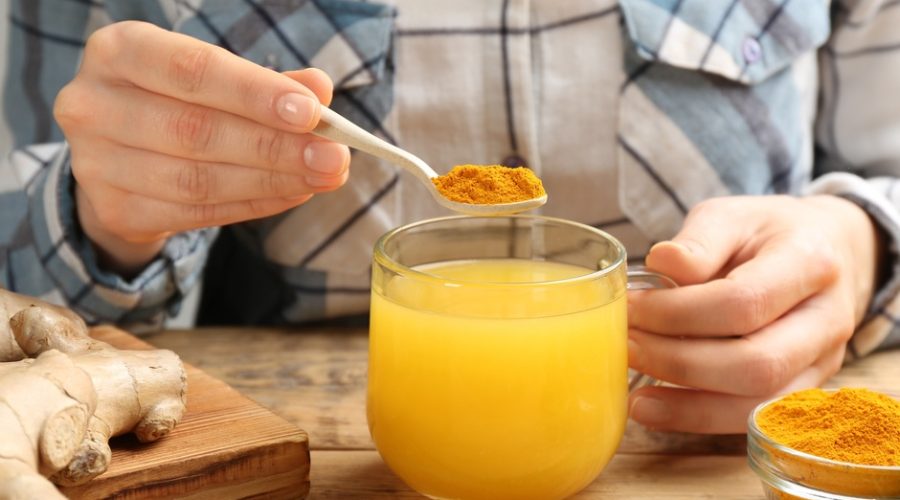 A woman adding a teaspoon of turmeric powder to a mug, up close. Oranges and ginger roots are also on the table.