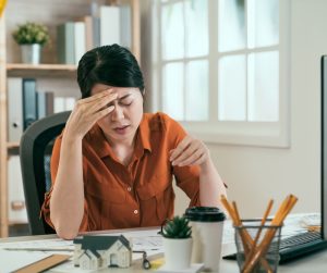 A woman sits at a desk with her head in her hands and a pained look on her face. A desktop computer sits in front of her.