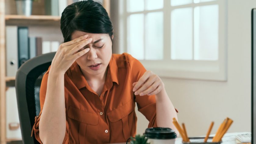 A woman sits at a desk with her head in her hands and a pained look on her face. A desktop computer sits in front of her.