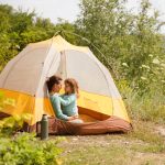 A woman and a young girl wearing matching blue shirts sit inside a yellow tent in a clearing with trees and wildflowers.