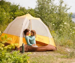 A woman and a young girl wearing matching blue shirts sit inside a yellow tent in a clearing with trees and wildflowers.