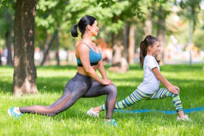 A mom and daughter work out together in a park on a sunny day. They exercise under the shade of trees.