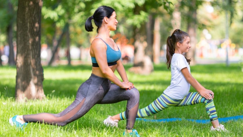 A mom and daughter work out together in a park on a sunny day. They exercise under the shade of trees.