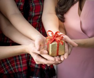 Three women holding a small present in front of them. It is wrapped in sparkly gold paper and a red ribbon.