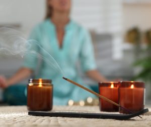 An incense smolders in a holder beside three burning candles. An out-of-focus woman performs a yoga pose in the back.