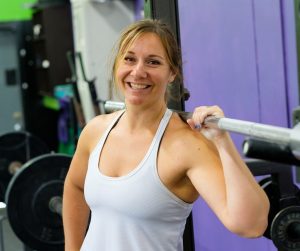 A fit and healthy mother at the squat rack in the gym smiling at the camera. She's in a tank top and has blue nail polish.