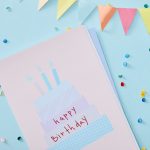 A birthday card with a cake reading "happy birthday" sits on a blue background with multicolored confetti and a banner.