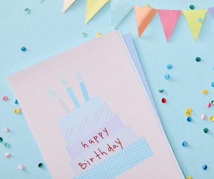 A birthday card with a cake reading "happy birthday" sits on a blue background with multicolored confetti and a banner.