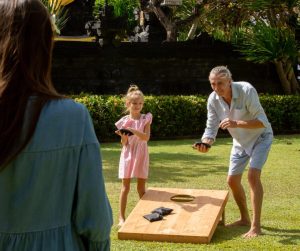 An older man and a young girl wearing a pink dress play cornhole outside on the grass as the sun shines.