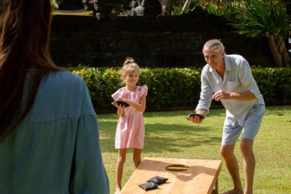 An older man and a young girl wearing a pink dress play cornhole outside on the grass as the sun shines.