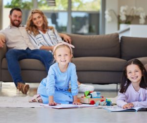 Two little girls sit on the floor with books and other toys. There are two adults sitting together on the couch behind them.