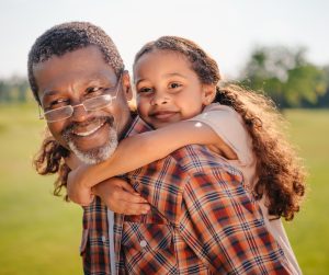 A smiling child with long hair looping their arms around their grandfather's shoulders as he carries them.