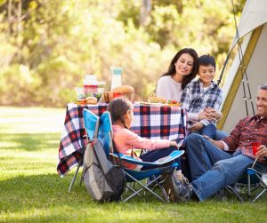 A family gathered in front of a tent, sitting on camp chairs around a small table with a checked tablecloth.