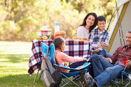 A family gathered in front of a tent, sitting on camp chairs around a small table with a checked tablecloth.