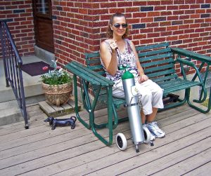 A woman in a tank top and capris sitting on a green bench on a patio. She is holding a portable oxygen tank on wheels.