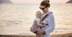 A woman stands on the beach looking at a baby she's carrying in a pink baby carrier. The baby faces the woman's chest.