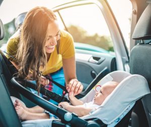 A smiling woman in a yellow shirt leaning into a car and playing with her baby, lying in their car seat.