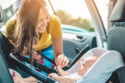 A smiling woman in a yellow shirt leaning into a car and playing with her baby, lying in their car seat.