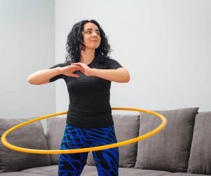 A smiling young woman spins a hula hoop around her waist. She works out in her living room in front of a gray sofa.