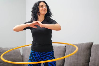 A smiling young woman spins a hula hoop around her waist. She works out in her living room in front of a gray sofa.